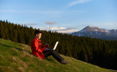 Shot of a cheerful young woman working on her laptop resting on top of the mountain on sunset after...