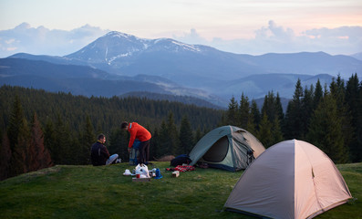 Male and female tourists in the camping near two tents while hiking together with their backpacks. On the background beautiful forests and mountains. Carpathians mountains, Ukraine