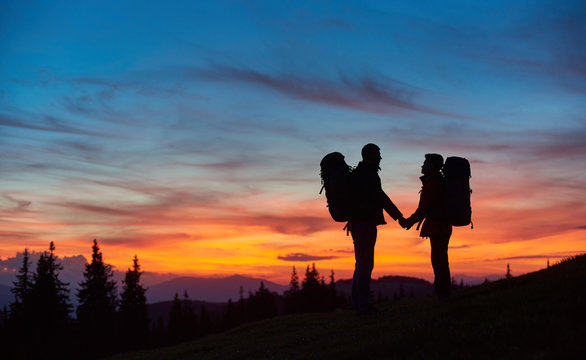 Loving couple tourists hiking together holding hands standing with their backpacks on top of a mountain during stunning sunset people love silhouettes meadow active relationships