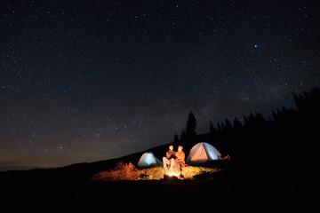 Night camping. Couple tourists sitting at a campfire near two illuminated tents under night starry sky. Long exposure