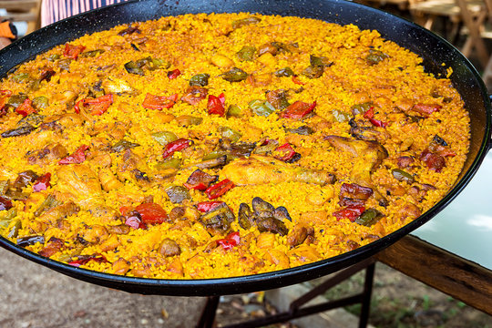 Large flat frying pan with cooked homemade Spanish paella with variety of meats, vegetables, rice, tomato sauce, spices. Outdoors, vibrant orane red and yellow colors.