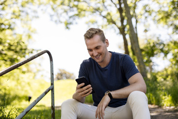 Handsome blonde man sitting down outdoors relaxed using his cellphone on a sunny summer day.