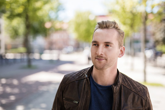 Satisfied man with a smile standing on walkway in the city wearing a brown leather jacket and blue tshirt on a sunny summer day.