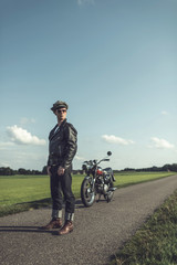 Vintage motorcyclist in leather jacket and cap standing with motorcycle on country road.