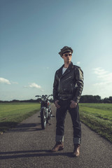 Vintage motorcyclist in leather jacket and cap standing with motorcycle on country road.