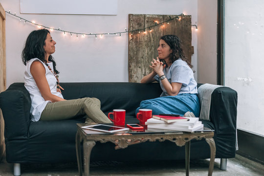 Two women friends talking to each other on a couch in apartment