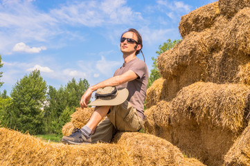 Naklejka na ściany i meble Handsome man with hat sitting on straw bales on a sunny day.
