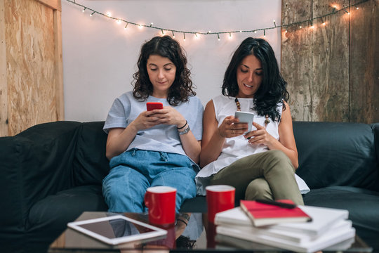 Two women friends use their smart phone in apartment on sofa