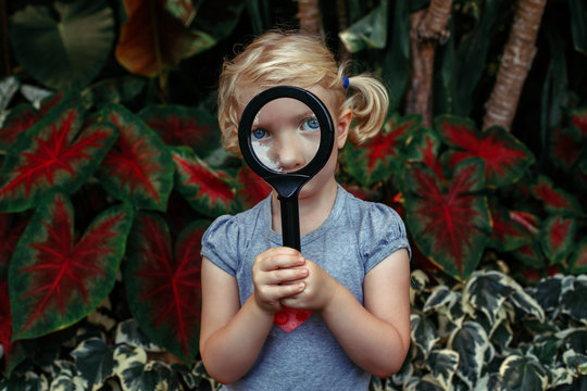 Portrait of cute adorable white Caucasian girl looking in camera through flowers through magnifying glass. Child with loupe studying learning nature. Early development education concept.