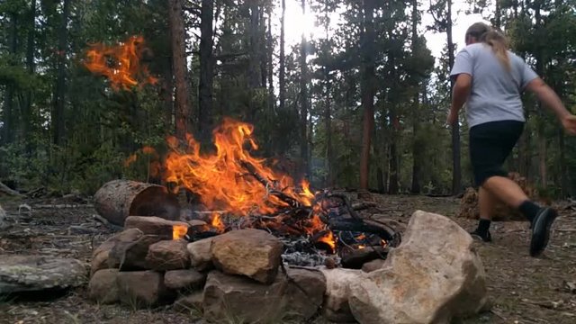 Burning Large Fire In Fire Pit In Pine Forest With Sun Shinning From Behind Trees And Running Man To Gather More Wood In Slow Motion. Slightly Windy Day Push Fire Toward Camera.