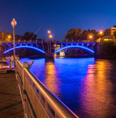 Night time image of river and bridge with blue lights, reflected in water with street lights. Anoka Minnesota