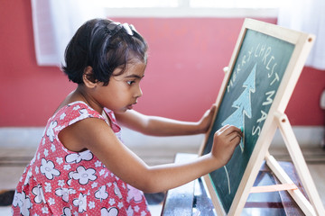 Little girl drawing Christmas tree on green chalkboard