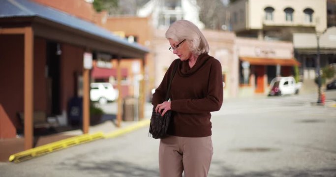 Lovely elder woman walking down street with purse pausing to answer text. Senior woman messaging on cell phone smiling and walking off camera outside. 4k