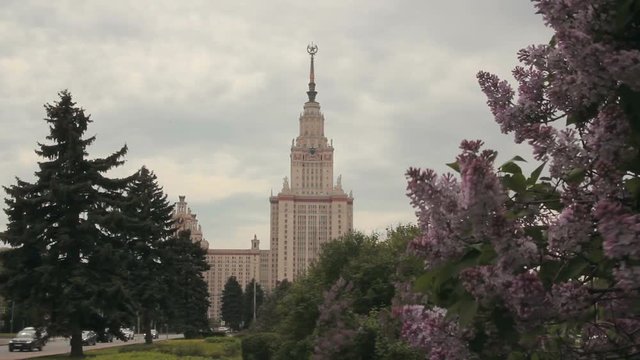Main building of Moscow State University (also known as MGU) behind lilac bushes on a cloudy day.