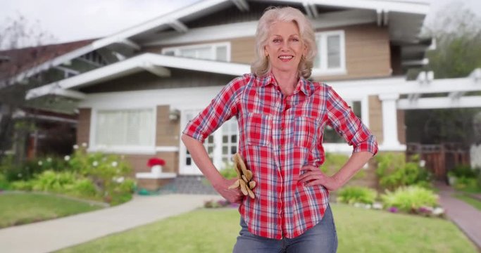 Portrait of proud senior woman standing with hands on hips outside home holding gardening gloves. Friendly elder woman in flannel shirt smiling at camera outside suburban house. 4k