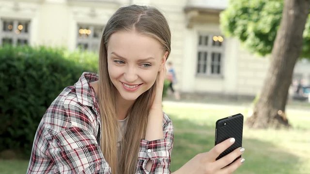 Pretty girl in checked shirt doing selfies on smartphone in the park, steadycam shot

