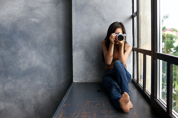 Picture of talented young unrecognizable brunette woman photograpther in jeans holding photo camera taking pictures or making setting, sitting barefooted on windowsill by large panoramic window