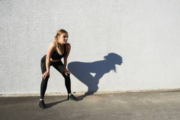 Picture of beautigul tired sporty young female with perfect athletic body in black sportswear and sneakers placing hands on her knees while having break after rmorning running workout outdoors