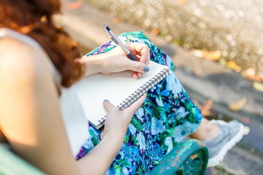 Close up left hand asian women with pen writing notebook and sitting on a wooden bench and a park.