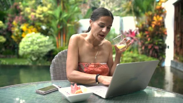 Attractive woman drinking juice and using laptop while sitting in exotic place, steadycam shot
