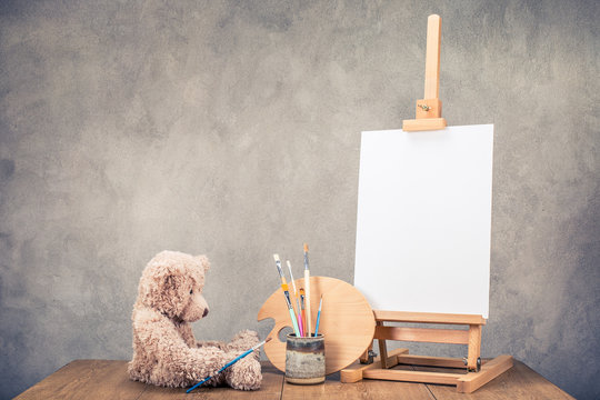 Teddy Bear toy, portable desk easel for painting with canvas blank, brushes and artist's palette on wooden table front concrete wall background. Retro instagram old style filtered photo