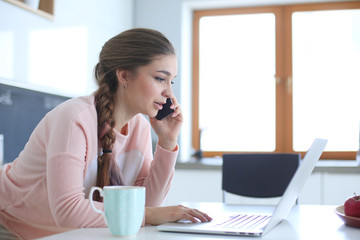 Young woman sits at the kitchen table using a laptop and talking on a cell phone.
