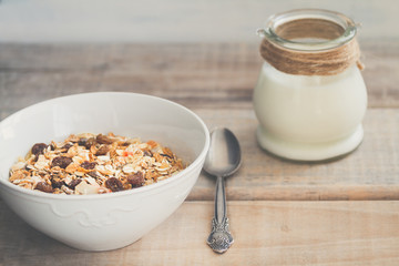 White bowl with Granola or Muesli and a jar of milk or plain yogurt and a spoon on a vintage wood background. Healthy breakfast composition.