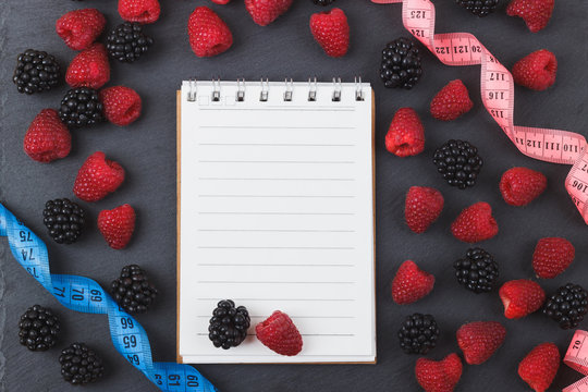 Red raspberries, blackberry, tape measure and notebook on the black slate stone background