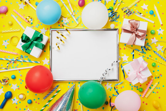 Party or birthday background. Silver frame with colorful balloon, gift box, carnival cap, confetti, candy and streamer on yellow table top view. 