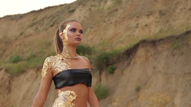 Attractive model with golden skin and piercing in her belly and professional makeup walking on the beach. Young blond model walking on the beach in a black leather swimsuit. Slowmotion shot