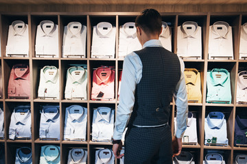 elegant man stands near a shelf with a lot of shirts and wants to buy one of them. Suite shop