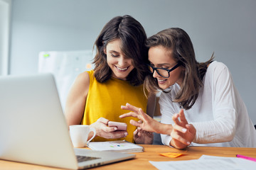 Two joyful coworkers browsing phone laughing together while taking break from work in the office