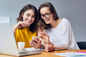 Couple of happy businesswomen looking at phone with laptop on desk in modern home office