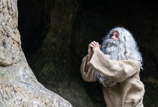 The Old Hermit Praying In The Cave. The Bearded Monk Meditating In Nature.