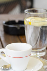 Close-up photo of a cup of coffee on a saucer with a spoon next to a glass of water and an ashtray