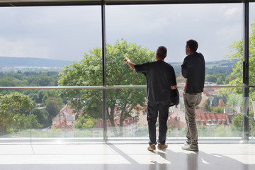 Two business man looking out of modern office window pointing with arm, from back