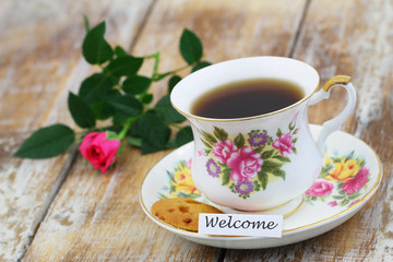 Welcome card with cup of tea in vintage porcelain up, cookie and pink rose
