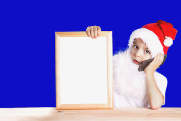 Young Santa Claus sitting at a wooden table and talking on the phone. Holding a blank picture frame with white background. Blue background. Chromakey. Close-up.