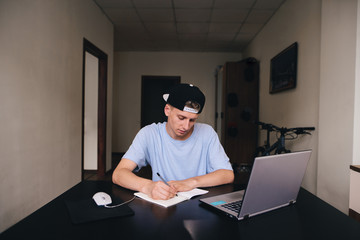 A teenage student writes a task in a notebook sitting at a table near the computer. Student studies at home.