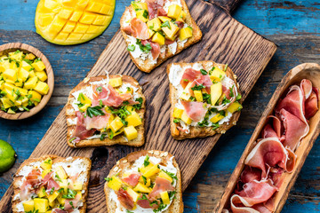 Toasts with cream cheese, ham jamon serrano and mango served on wooden board with red wine, blue wooden rustic background, top view