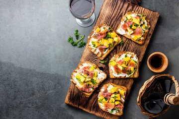 Toasts with mango, cream cheese, ham jamon serrano served on wooden board with red wine, gray slate background, top view. Copy space