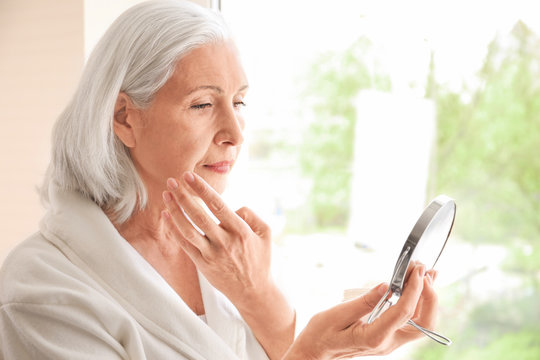 Beautiful Elderly Woman Holding Mirror And Applying Face Cream At Home