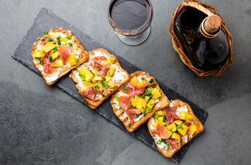 Toasts with cream cheese, ham jamon serrano and mango served on black slate board with red wine, top view, gray slate background