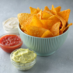 Crispy mexican snacks nachos or tortilla in bowl with dipping sauces on gray table.