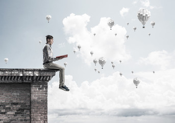 Man on roof edge reading book and aerostats flying in sky