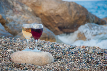 Two wineglasses with red, white drinks stay on grey stone on sea beach with blue waves and pebble