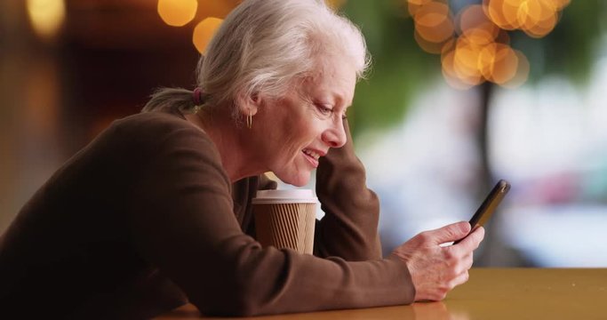 Slowmo of mature Caucasian woman sitting at table with coffee texting on cellphone. Cheerful senior woman messaging on smart phone in outdoor setting smiling and laughing. 4k 