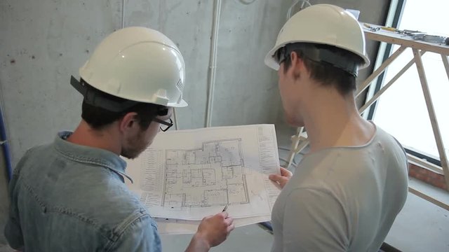 Master gives instructions according to the drawing on the paper. Two men in white protective helmets on their heads discuss the upcoming work to be done according to the scheme. Project Manager with