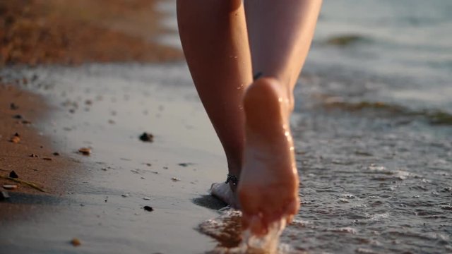 Legs of caucasian girl wearing shorts and walking barefoot wet sand sea beach, sun, lens, flare. Slow motion. Woman walks away from the camera