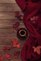 Knitted scarf of burgundy color with autumn leaves and a cup of coffee on a dark wooden background.